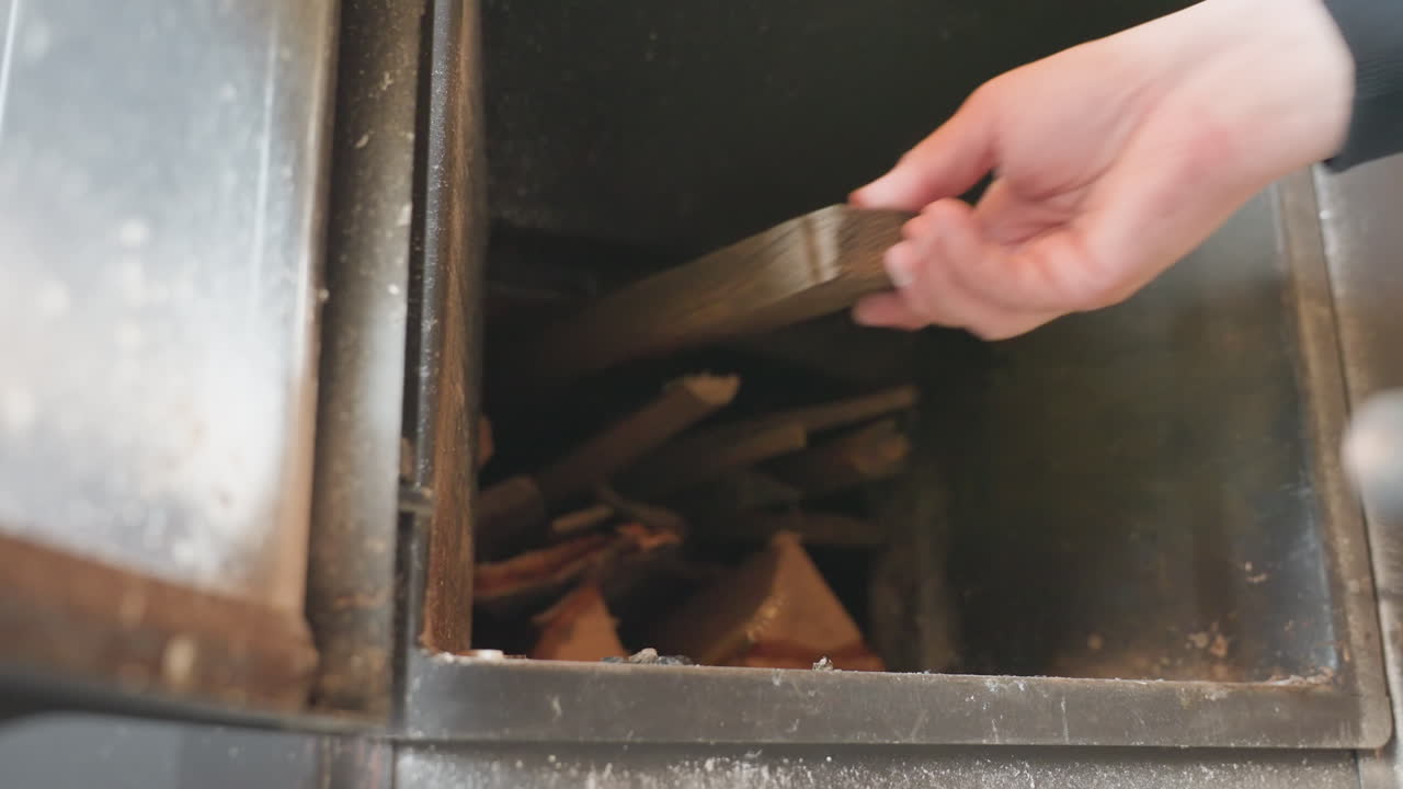 Extreme close up through iron stove door shows person hand arranging split fire wood inside dark chimney box, stacking kindling for ignition, promising cozy interior warmth during cold evening