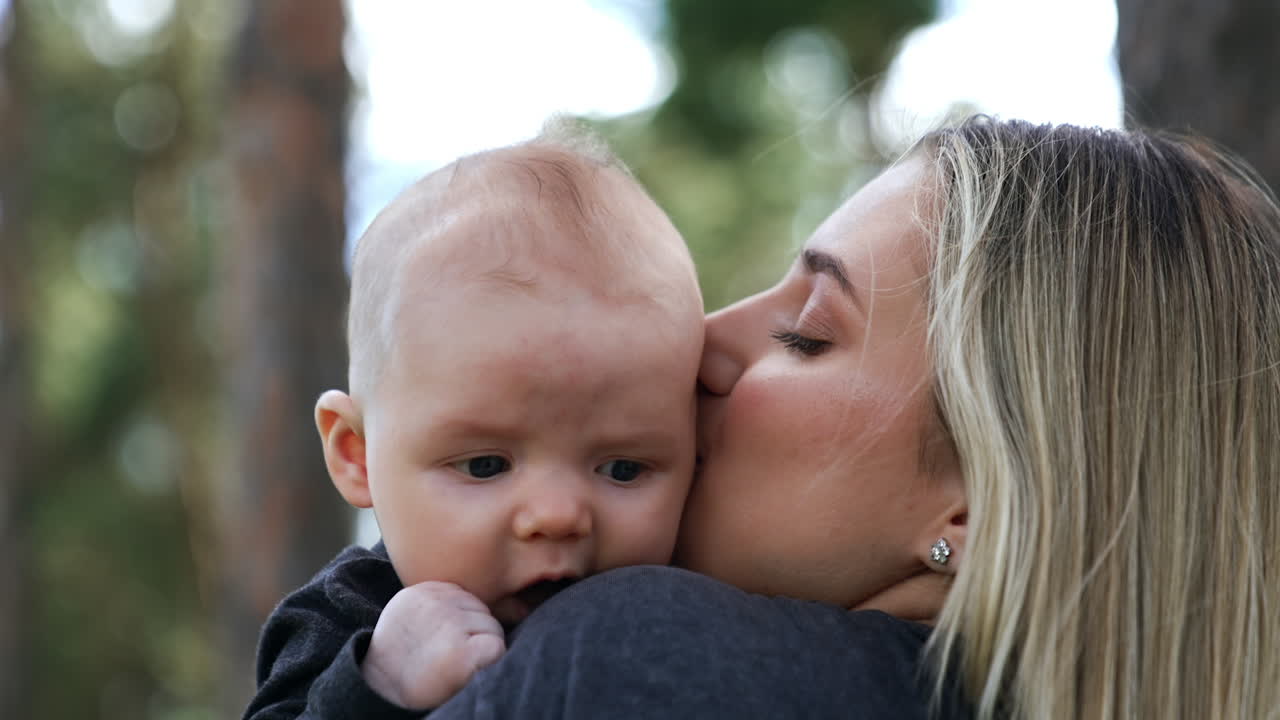 Cute little newborn rested his head on mom's shoulder. Loving mom kisses her adorable son on the ear. Blurred backdrop.