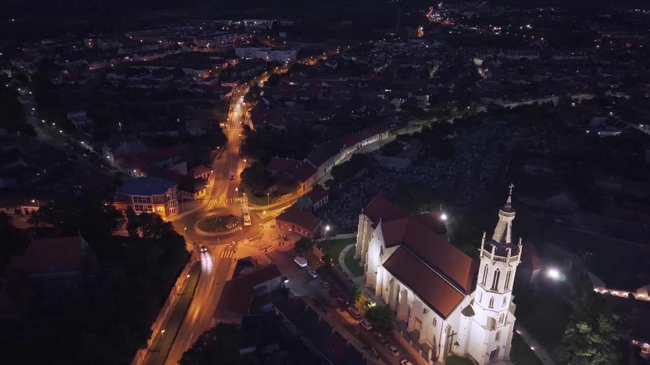 A stunning aerial night view of a beautifully illuminated Gothic-style church in the heart of a historic town. Drone moving forward between the church and the roundabout