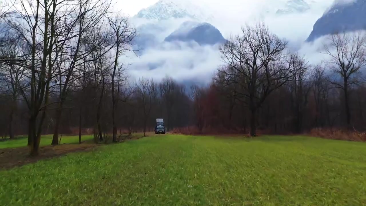 Un drone si avvicina a un veicolo a energia solare su una strada di valle, inclinando verso l'alto verso il Monte Ornavasso avvolto in nuvole basse e nebbia, rivelando cime alpine innevate e alberi invernali.