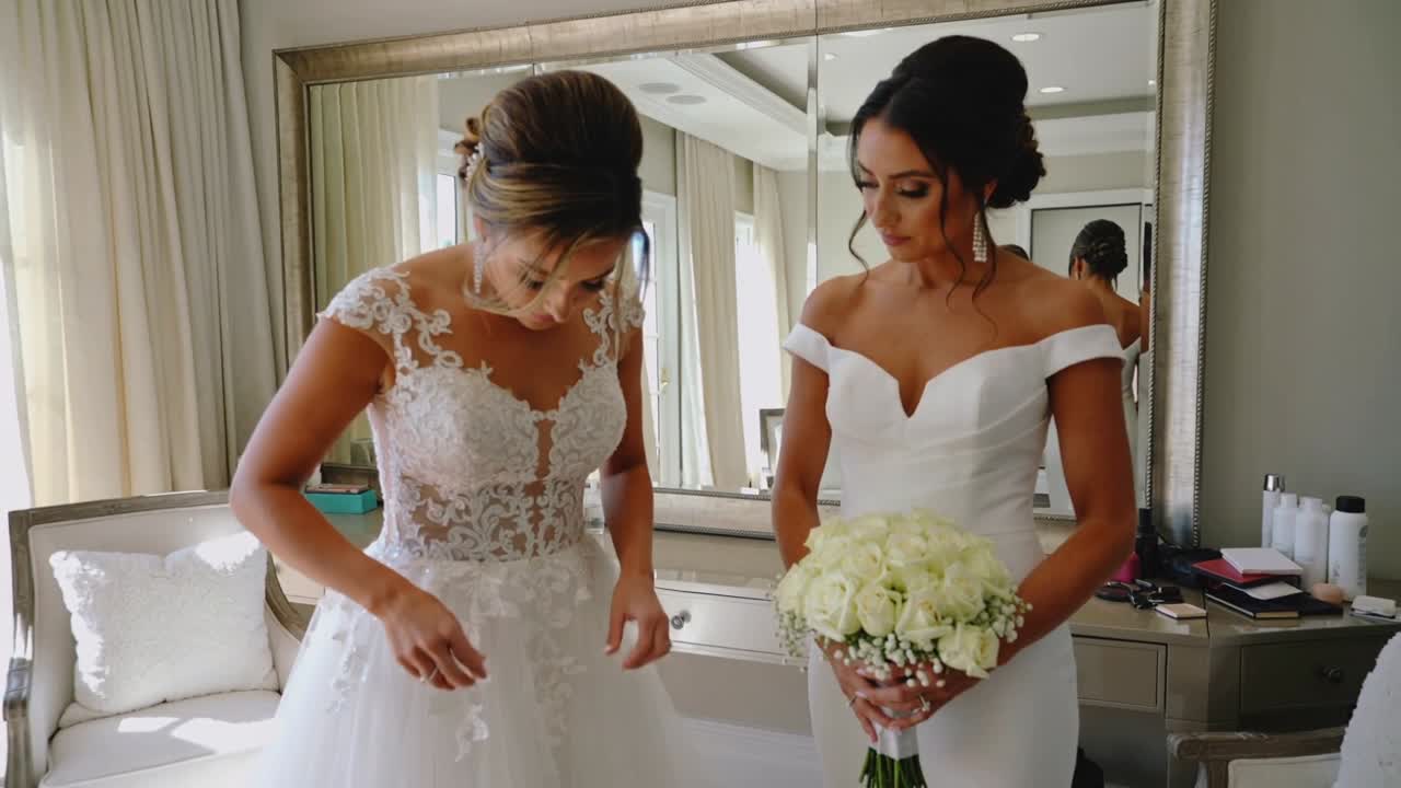 Adjusting bride fixing lace ballgown in bridal suite during prep, bridesmaid holding white roses