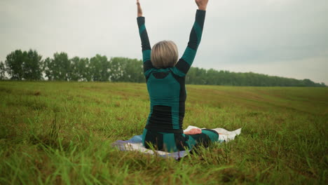 back view of woman in green and black suit seated on yoga mat with legs crossed, hands raised above her head, practicing yoga in a grassy field, yellow boots visible in background under cloudy sky