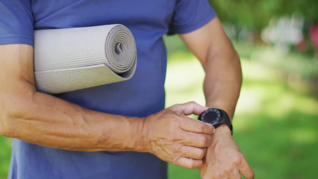 Video of midsection of biracial senior man holding yoga mat and checking smartwatch
