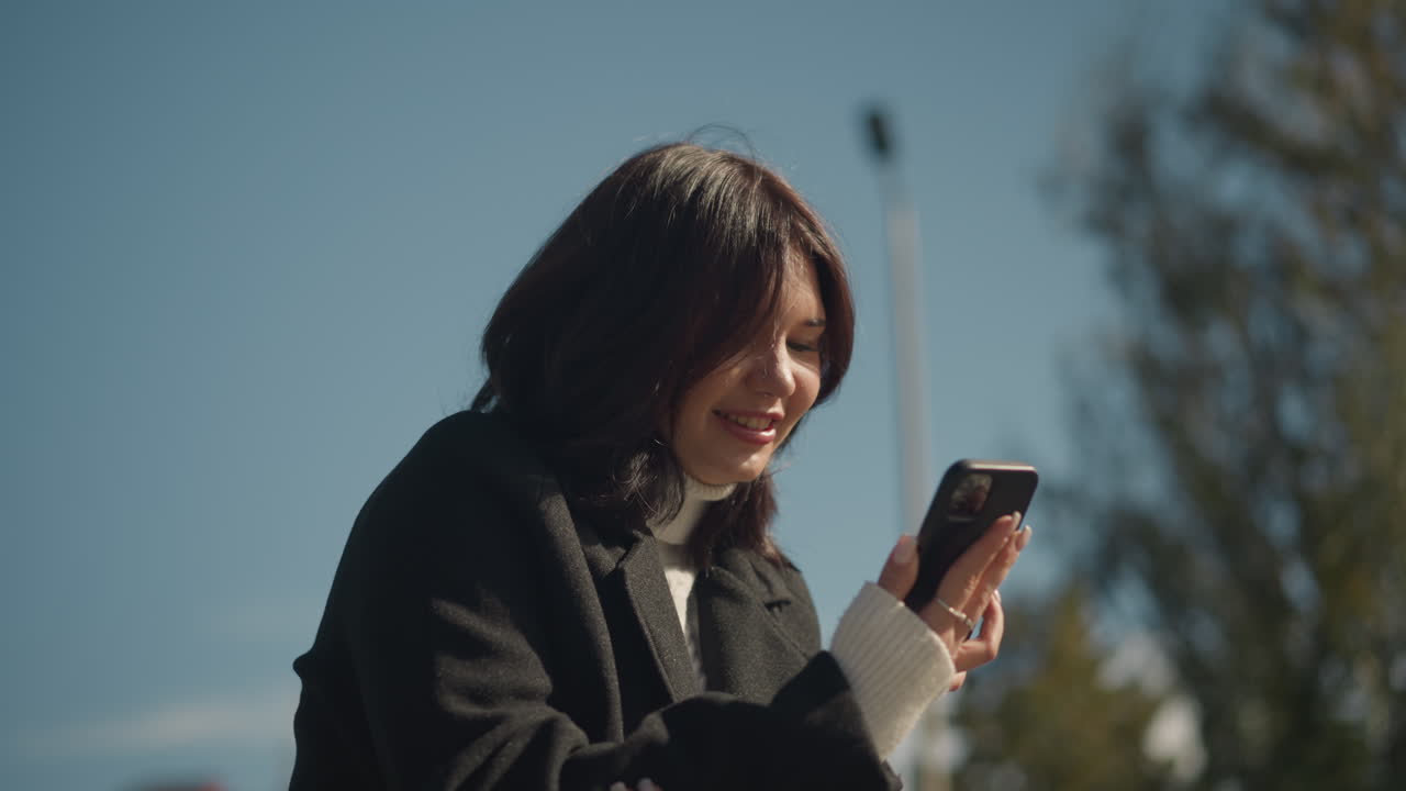 웃는 즐거운 여성, 밝은 은 날 배경에서 부드럽게 흔들리는 흐릿한 초록색과 함께 공립 공원에서 그녀의 전화에 초점을 맞추는 동안 그녀의 머리카락을 조정