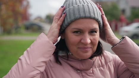 happy woman wearing pink jacket and grey beanie adjusting beanie with a warm smile, blurred background featuring buildings and parked cars