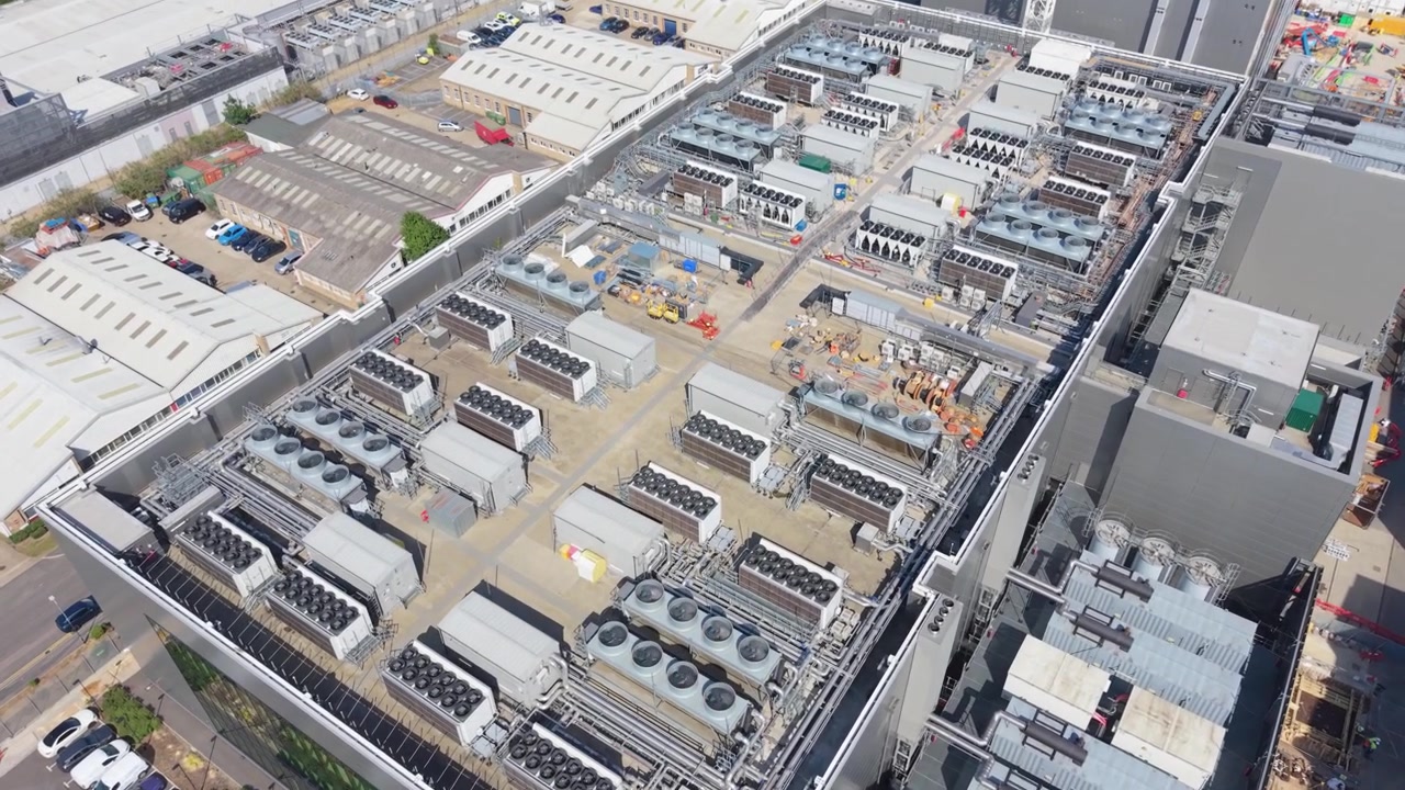 Una vasta prospettiva aerea mostra una grande struttura di data center con più edifici e ampie unità di raffreddamento. Osservare l'intricata infrastruttura che opera sotto cieli diurni sereni