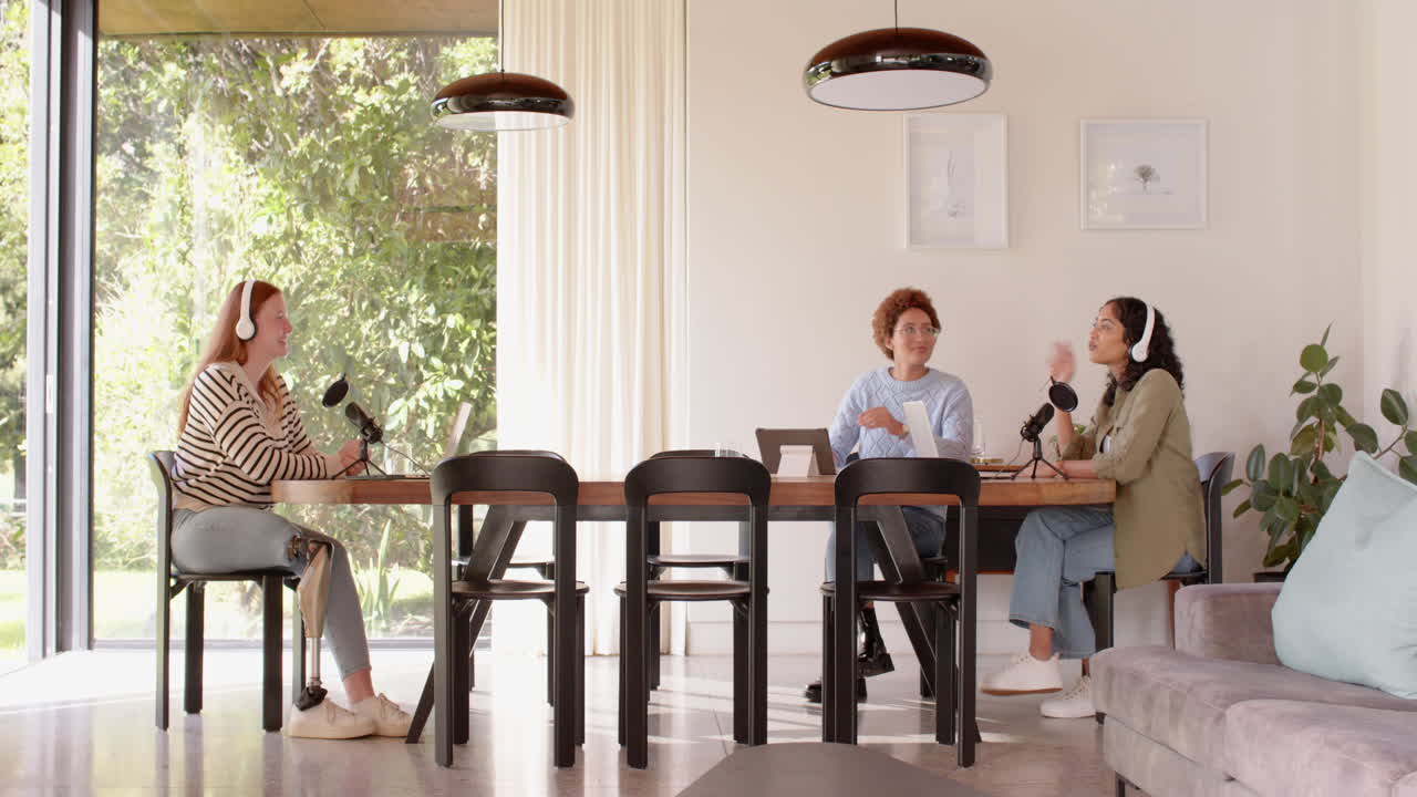 Recording podcast, diverse female friends sitting at table with microphones and laptops