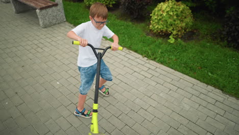 a young boy spins his scooter in a playful clockwise direction on a well-maintained interlocked path in a park, the lush greenery and nearby bench