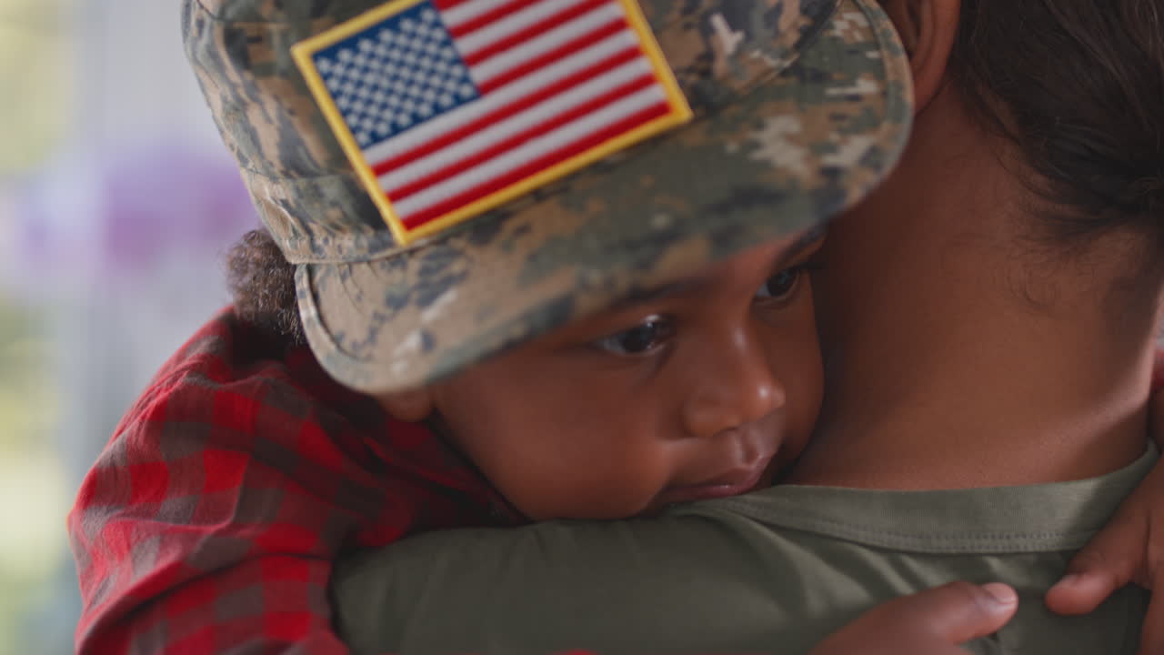 Madre militare americana in uniforme a casa in licenza che abbraccia il figlio che indossa il suo berretto nella cucina di famiglia.