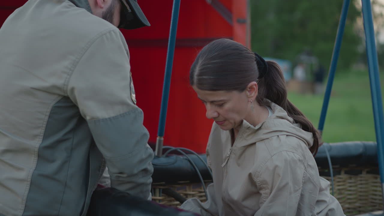 man hands gas cylinder to woman preparing burner assembly inside wicker balloon basket during preflight setup on grassy roadside under cloudy sky with red envelope backdrop