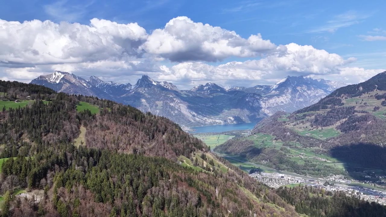 Ampia panoramica aerea di un lago di montagna circondato da valli alpine, foreste e cime spettacolari nelle Alpi svizzere. Paesaggio primaverile scenografico sotto formazioni nuvolose dinamiche