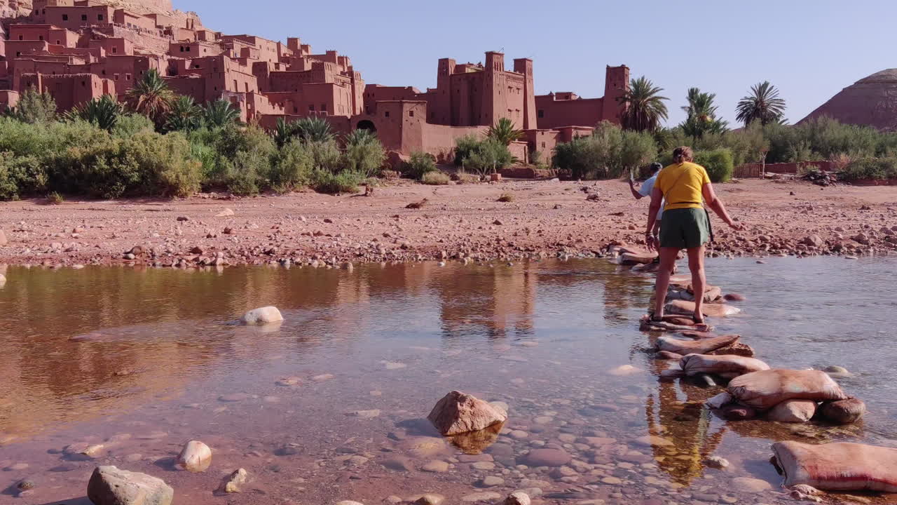 Couple Crossing The River via Stepping Stones Near Ait Benhaddou Historic Kasbah (fortified village) In Morocco. Static Shot