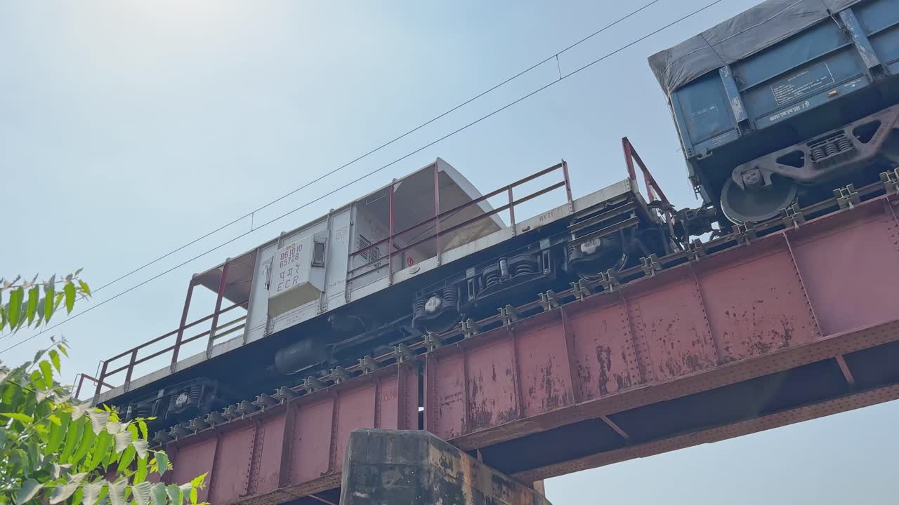 A moving freight train reveals its guard cabin as the camera pans beneath a red steel bridge, showcasing wheels, railings, and rugged industrial details
