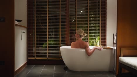 detrás de una mujer rubia desnuda en la bañera blanca del resort, disfrutando sola de un baño caliente