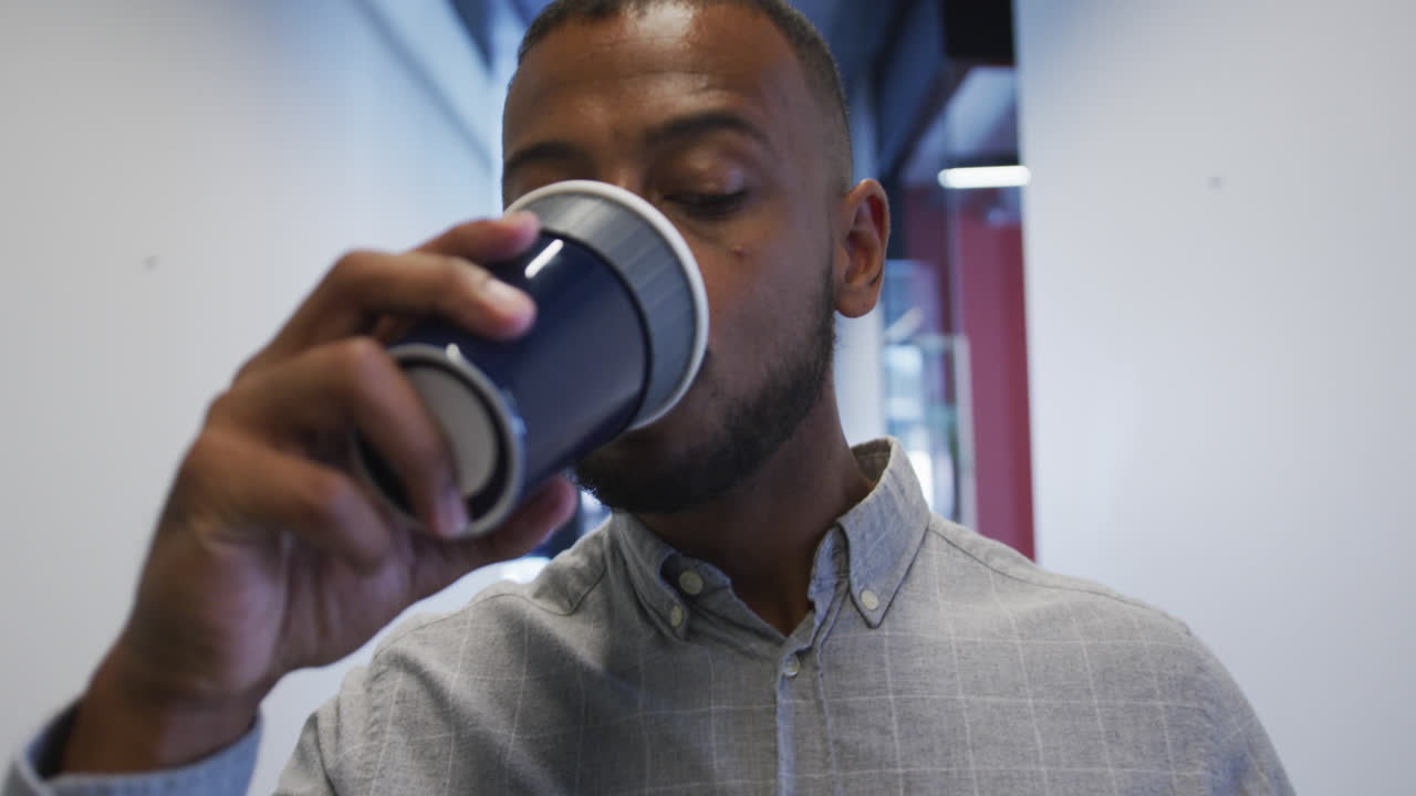 hombre de negocios biracial caminando y bebiendo una taza de café en una oficina moderna