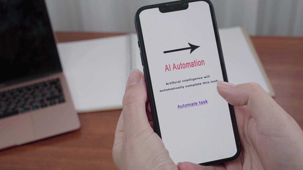 Hands hold a smartphone showing an AI Automation message with an Automate task link above a laptop on a wooden desk.