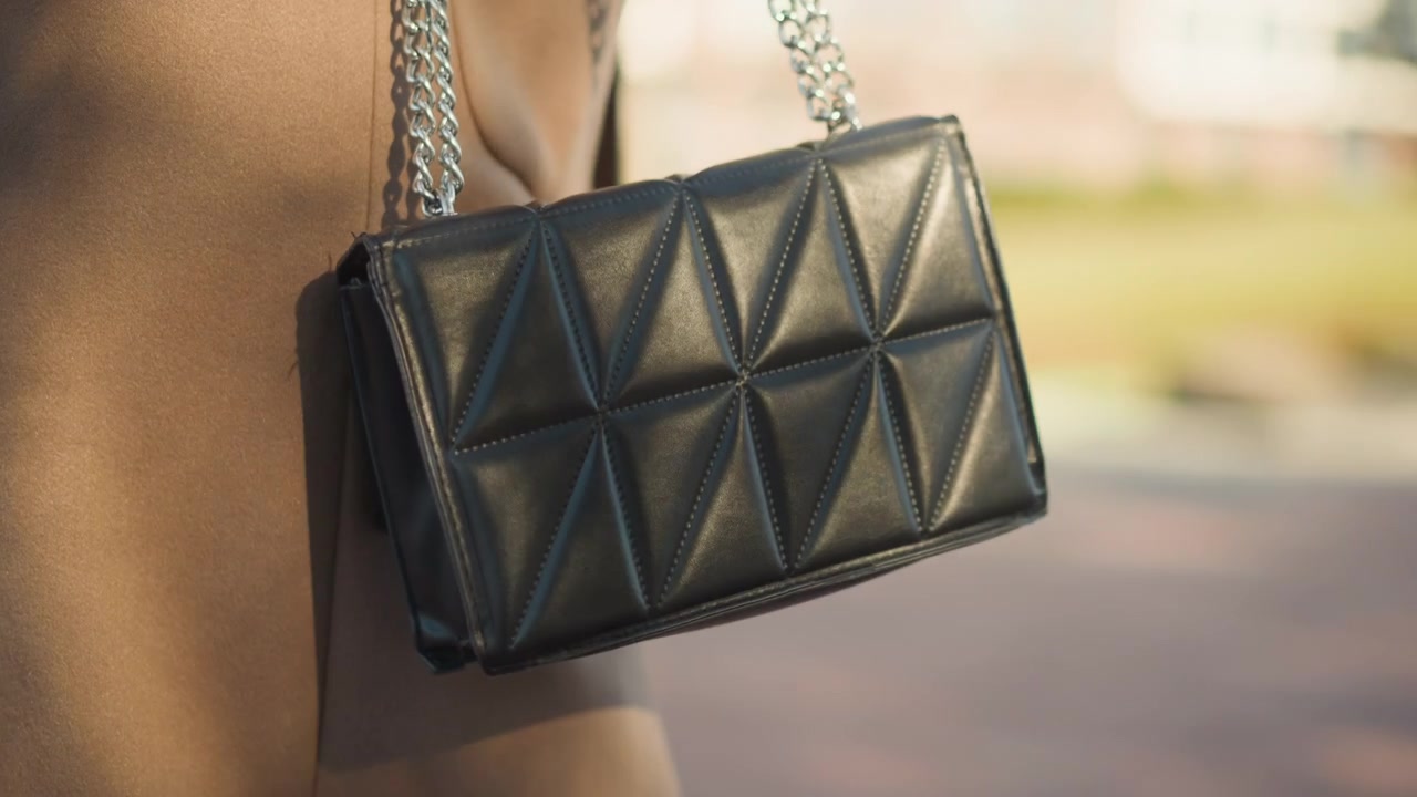 Primo piano di una borsa a tracolla in pelle trapuntata nera con tracolla a catena argentata appesa alla spalla di una donna. La borsa presenta cuciture geometriche e riflessi di luce soffusa su uno sfondo esterno sfocato.