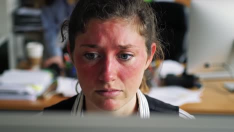 An employee in a modern office concentrates on her work while surrounded by desks, paperwork, and other staff engaged in similar tasks. The atmosphere appears intense yet collaborative.