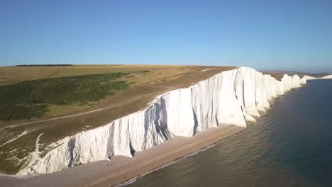 Vista-De-Drones-De-Acantilados-En-La-Costa-Del-Reino-Unido-Con-Mar-Y-Playa-De-Arena-Aves-Pov-Movimiento-Suave-Naturaleza-Diferentes-Tonos-De-Color-De-Vegetación-Cielo-Azul-Claro-Documento-De-Película-Cinematográfica-Lenta