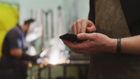 Trabajador-De-Fábrica-Caucásico-En-Una-Fábrica-De-Pie-En-Un-Taller,-Usando-Su-Teléfono-Inteligente