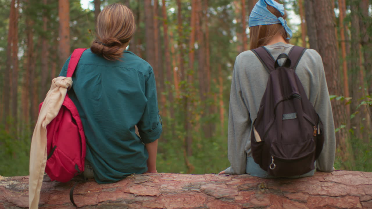 Back view of hikers sitting on fallen tree in lush forest, surrounded by tall trees, one rests hand on tree with long nails, while companion in green shirt relaxes nearby