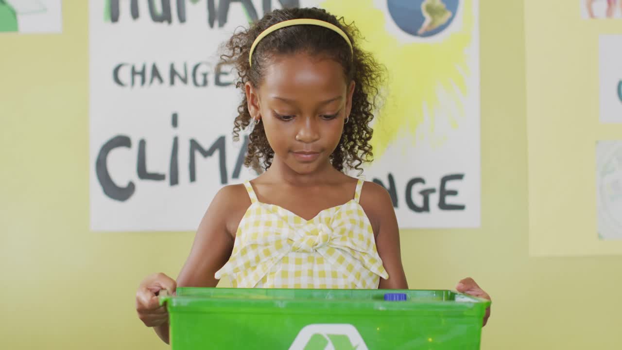 Video of happy african american girl holding box with recycling symbol in classroom