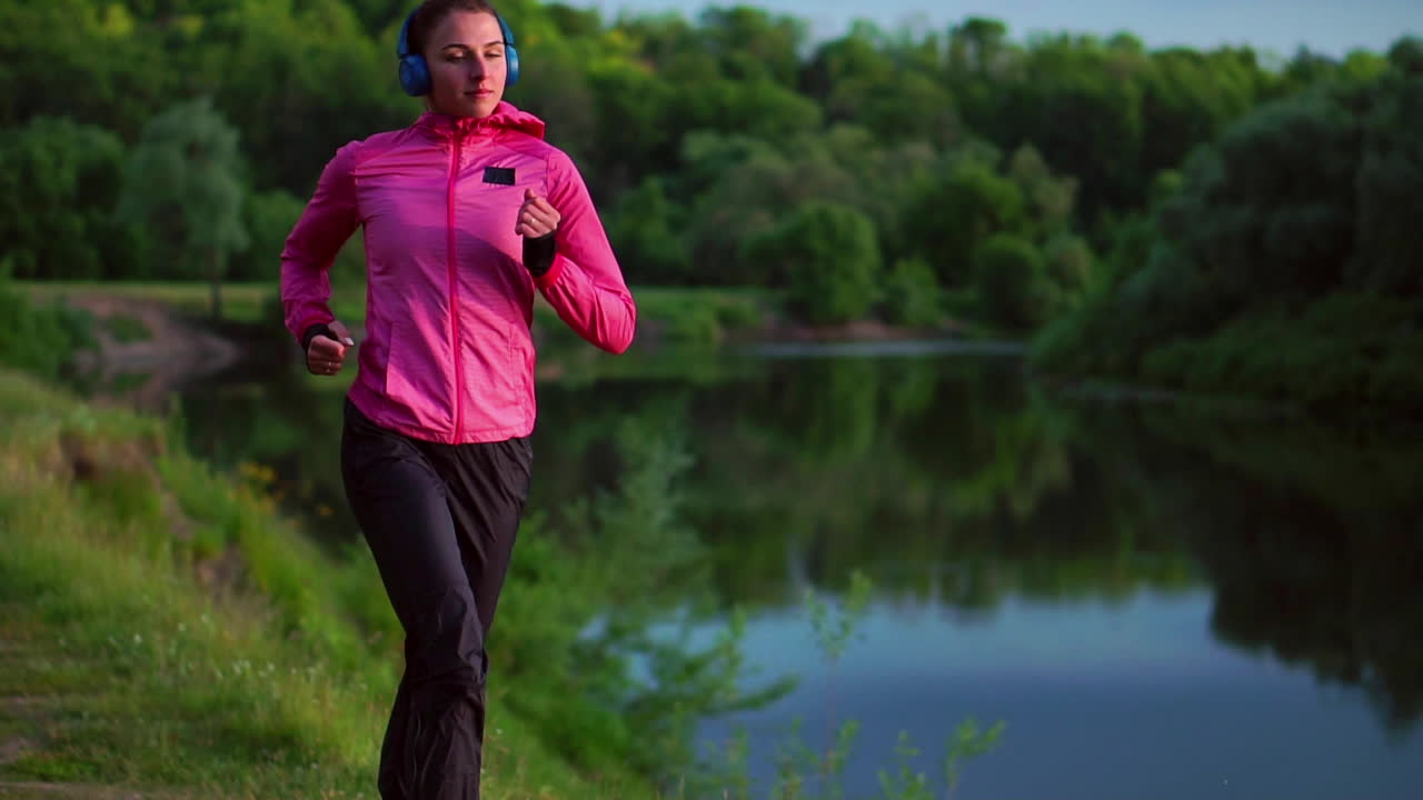 Brunette with long hair in headphones runs along the river in the Park in the morning at sunrise in the summer in a pink jacket and black pants