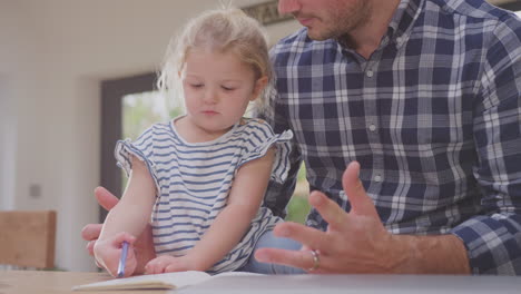 Father-at-home-at-kitchen-counter-helping-young-daughter-to-draw-picture-in-book---shot-in-slow-motion