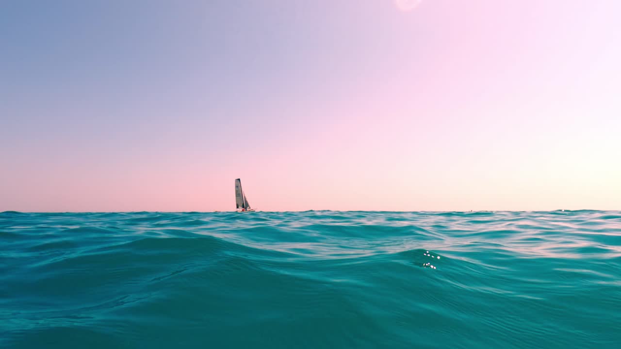 vista en ángulo bajo del nivel del mar de un pequeño velero navegando en aguas tranquilas y abiertas de color turquesa con cielo rosa en el fondo