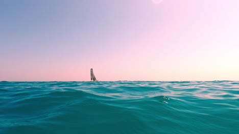 low-angle sea-level view of small sailboat sailing in calm open turquoise sea water with pink sky in background