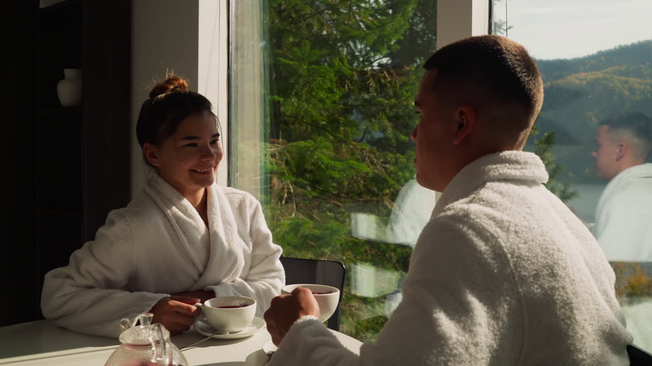 Couple talks drinking tea by window. Young people in bathrobes enjoy weekend drinking hot beverage in riverside hotel. Concept of vacation relaxation