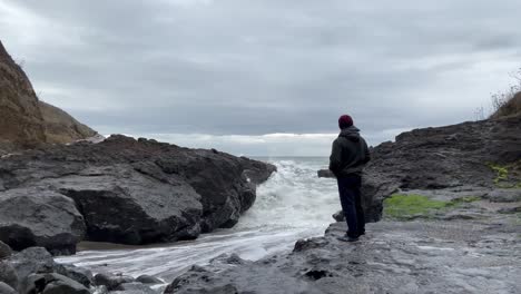 Lonely-Man-At-The-Rocky-Shore-With-Breaking-Sea-Waves-During-Cloudy-Day