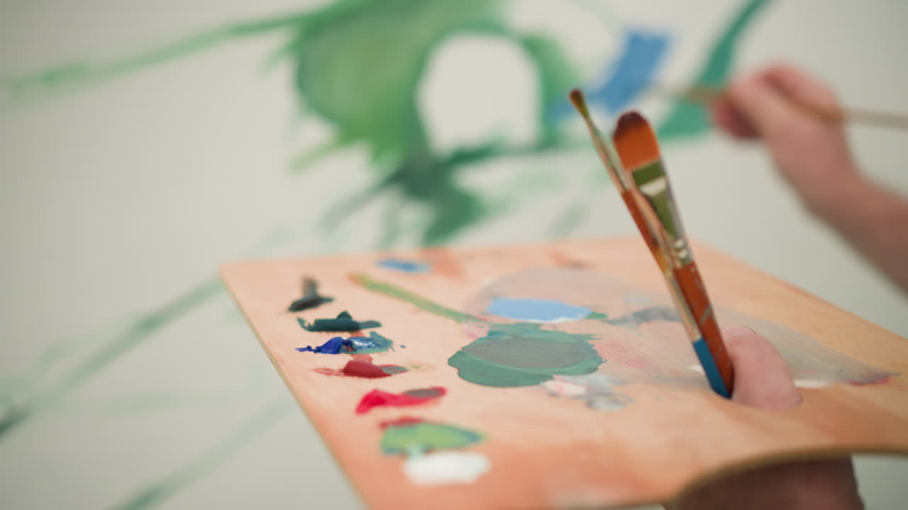 얼굴이 보이지 않는 화가 한 명은 다양한 활기찬 페인트 색으로 가득 찬 나무 패을 들고 있습니다. 체크색 셔츠를 입고, 예술가는 큰 캔버스에 페인트를 적용하는 데 집중합니다.