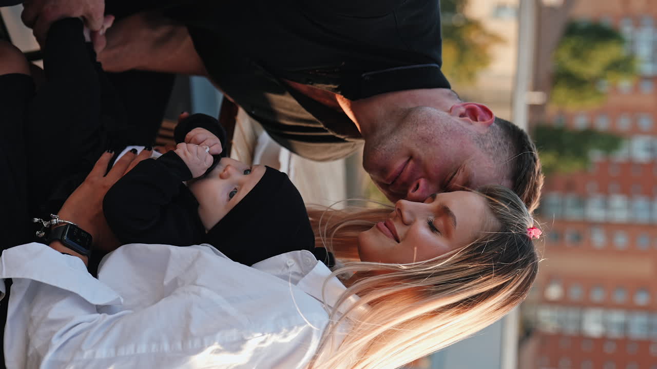 Blonde woman wearing white shirt holds her baby. Loving husband kisses his wife on the cheek. Vertical screen. Blurred city at backdrop.