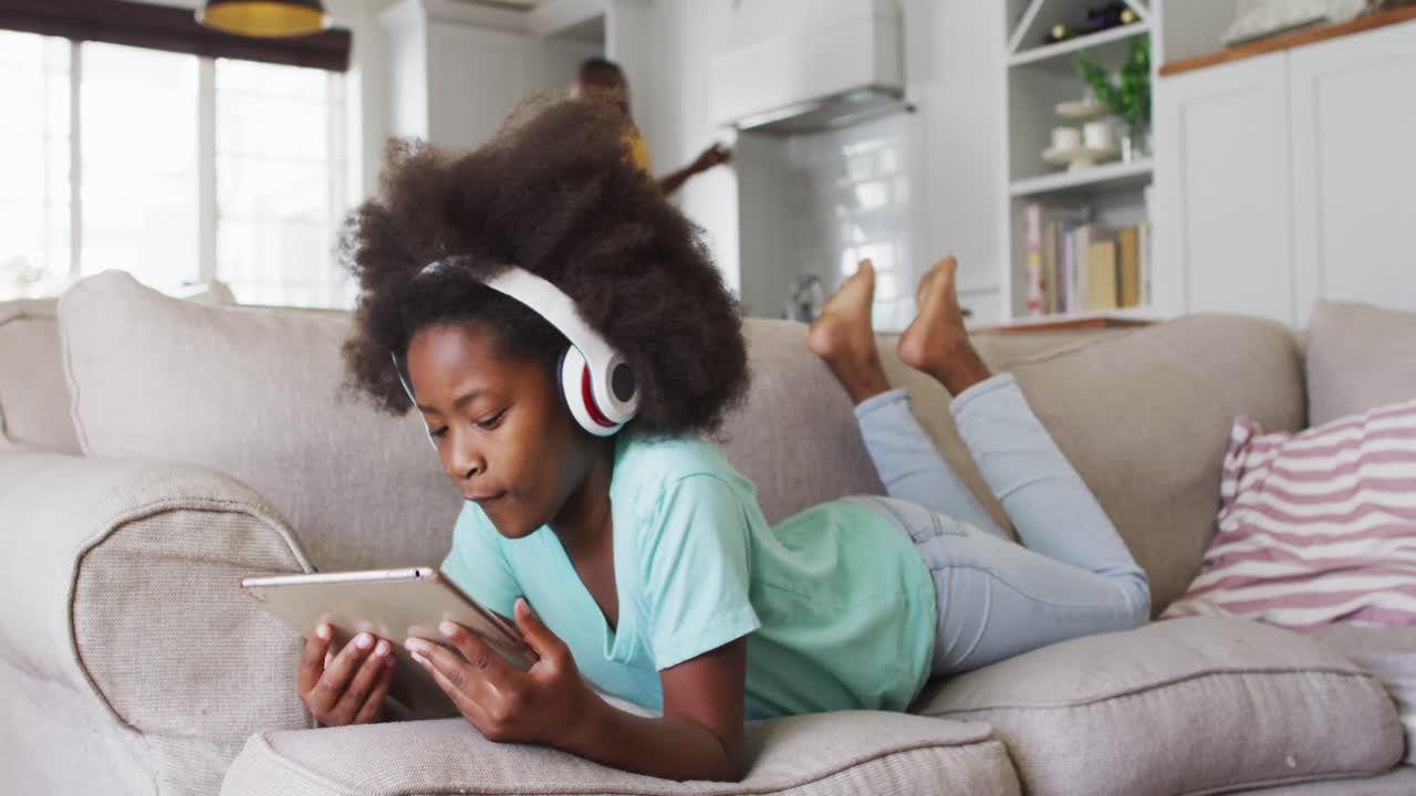 Ragazza afroamericana con le cuffie che usa un tablet sdraiata sul divano.