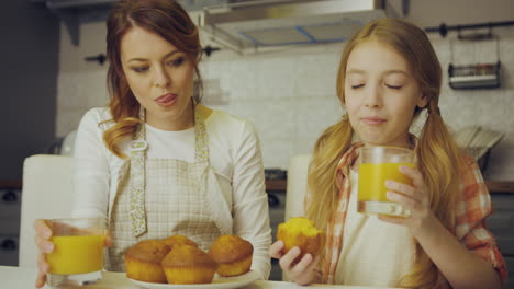 portrait shot of the attractive mother and her pretty teen daughter sitting in kitchen, eating muffins, drinking juice and hugging. indoor