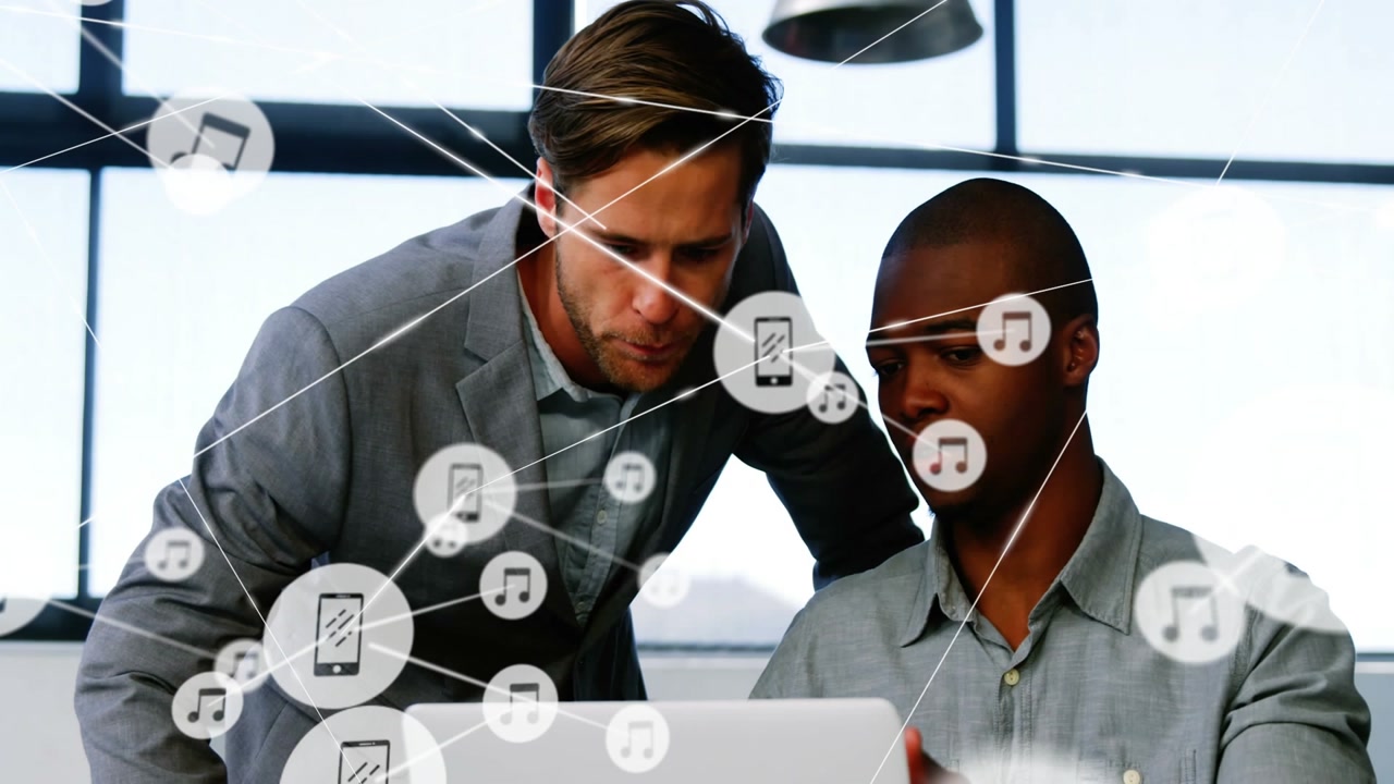 Seated colleague tapping laptop, standing lead leaning, pointing as music web expanding for review