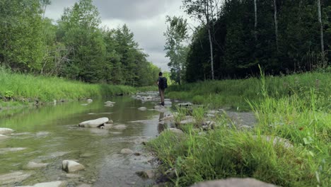 Single-lone-hiker-searches-a-river-bed-while-standing-on-a-rock-in-the-middle-of-a-river-flowing-through-a-forest-landscape