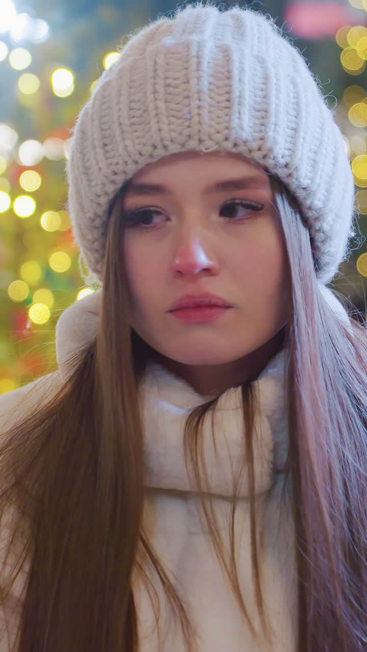 Young woman in cozy winter outfit sits outdoors at night, looking pensive, festive city lights illuminate the background as people skate, socialize, and enjoy the lively winter atmosphere around her