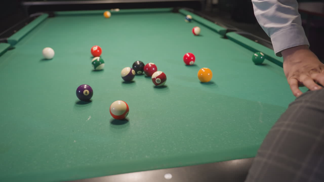 Close up of person sitting on green pool table using hand to roll orange billiard ball. Pool table covered with scattered colorful balls in dimly lit billiard room with relaxed ambiance