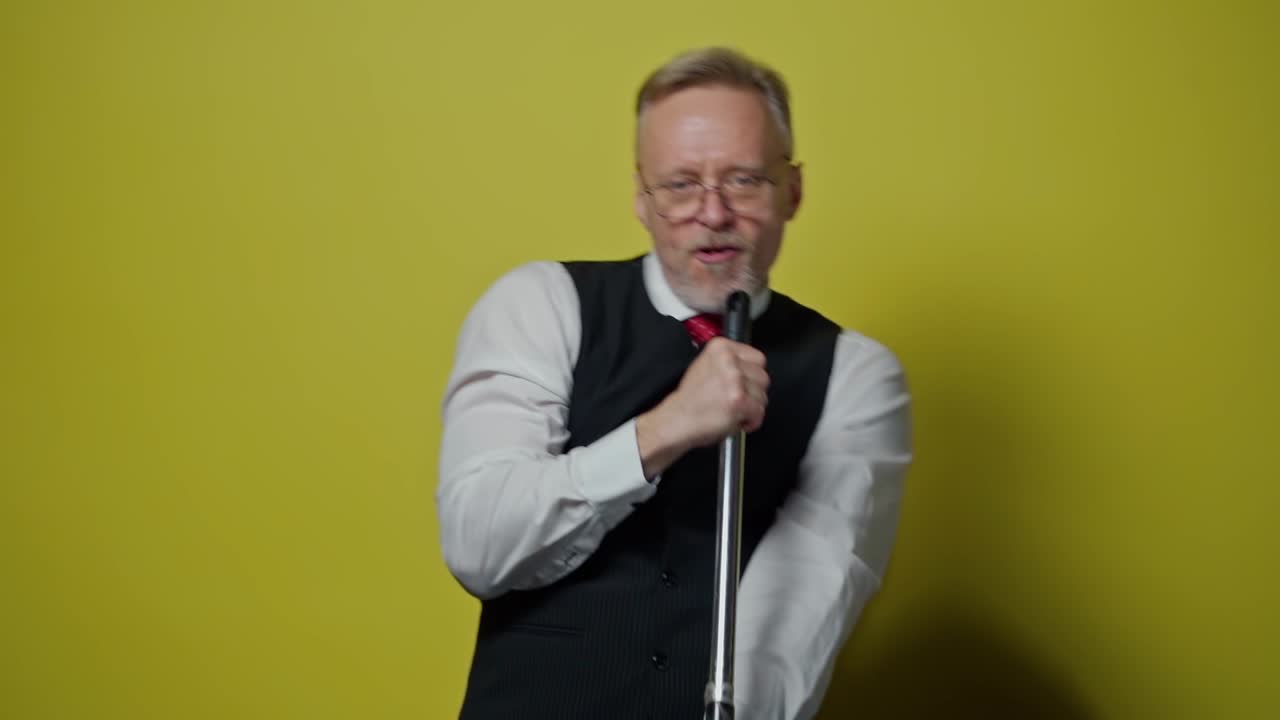 Senior positive man is having fun in studio. Funny mature man singing into a mop on the yellow background. Positive emotions.