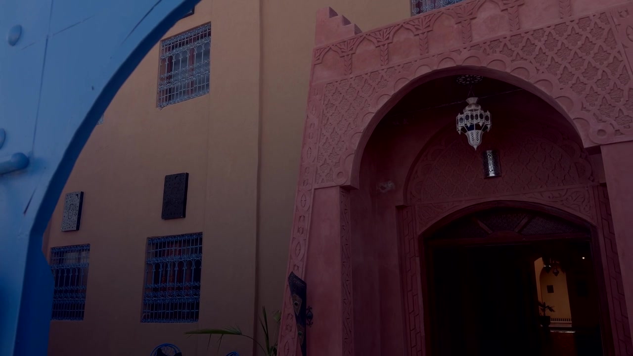 Carved red plaster mosque arch with decorative lantern beside a beige building with barred windows in Morocco