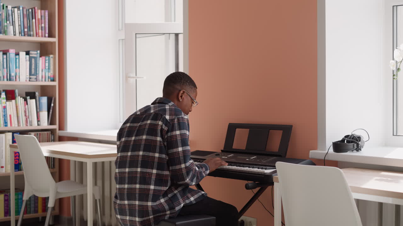 hombre con gafas disfruta tocando sintetizador. intérprete negro ensaya secuencia melódica en instrumento eléctrico junto a la estantería. ensayo en casa