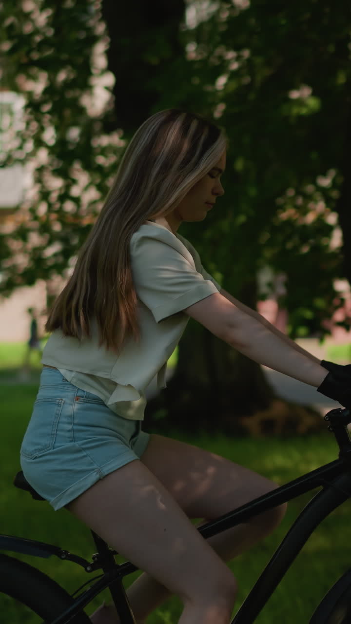 나무 가지로 부분적으로 숨겨진 울창한 초록색 공원을 통해 자전거를 자신감 있게 타고 있는 젊은 여성의 측면 시각, 긴 머리카락이 자유롭게 흐르고, 배경에는 초록색 잎자루와 건물이 있습니다.