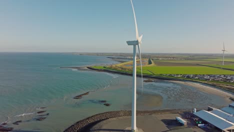 Toma-Aérea-En-Cámara-Lenta-De-Una-Turbina-Eólica-A-Lo-Largo-De-La-Costa-En-Una-Zona-Rural-En-Los-Países-Bajos-Contra-Un-Cielo-Azul-En-Un-Día-Soleado