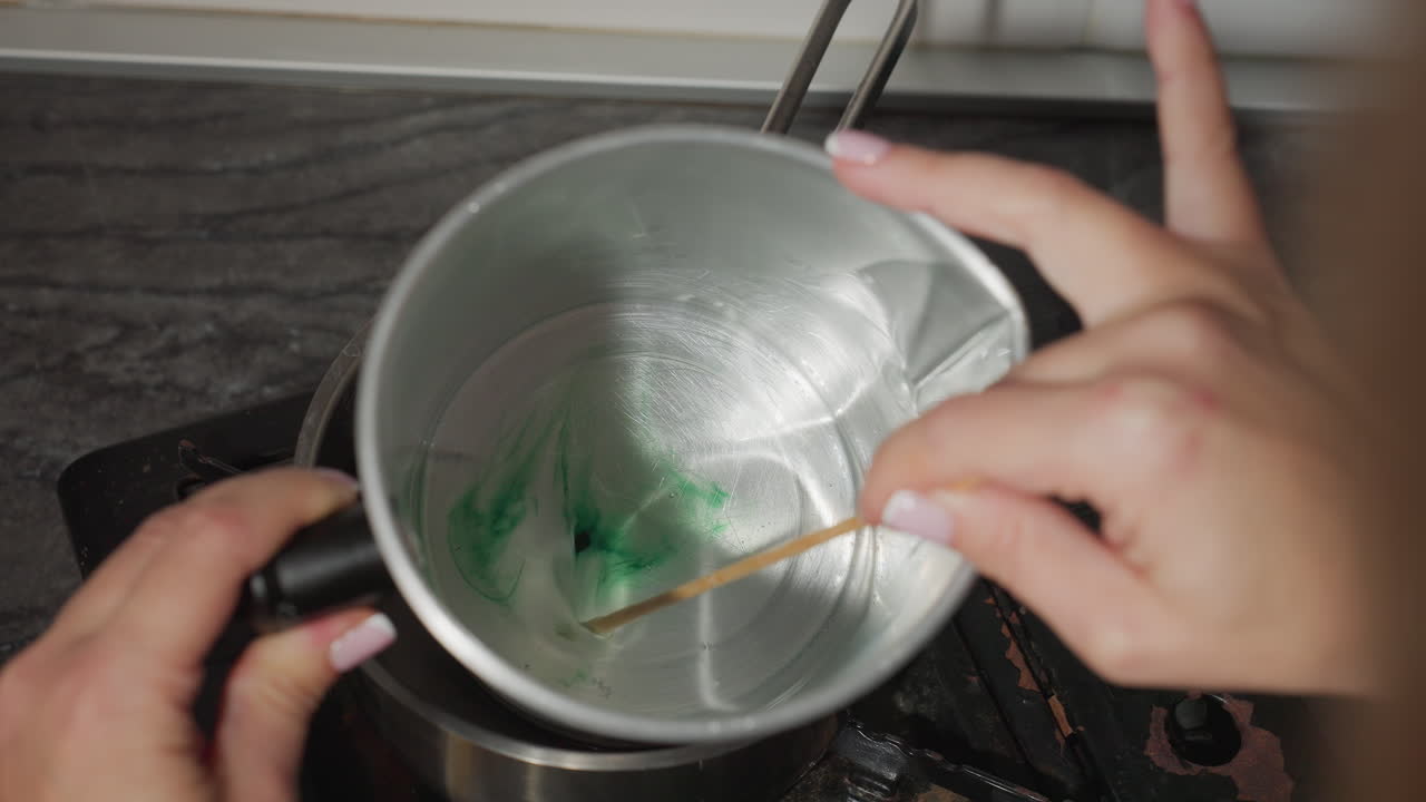 Close up of person stirring green liquid inside metal container placed in steaming pot on gas burner during craft preparation process in kitchen, focusing on melting and mixing colorful ingredients