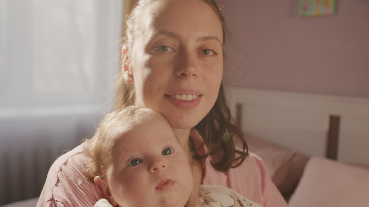 Porträt einer glücklichen, jungen Mutter, die ihr Baby in einem sonnendurchfluteten, gemütlichen Schlafzimmer hält