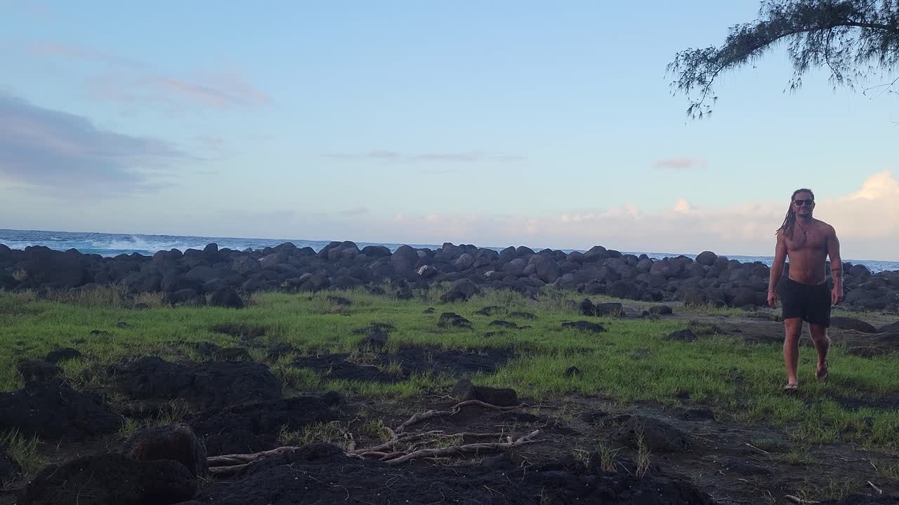 A shirtless man strolls across a lava-strewn grassy coastline at sunrise, approaching the camera in a peaceful tropical setting with distant ocean waves.