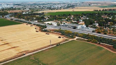 Ein-Nach-Oben-Fliegender-Drohnenschuss,-Ein-Riesiges-Gelbgrünes-Feld,-Mit-Auf-Einer-Straße-Fahrenden-Autos-Und-Einer-Stadt-Voller-Gebäude-Im-Hintergrund,-4K-Video