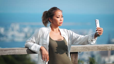 asian woman with dark blonde hair in a ponytail takes a selfie with a smartphone, leaning against a wooden railing with a cityscape in the background